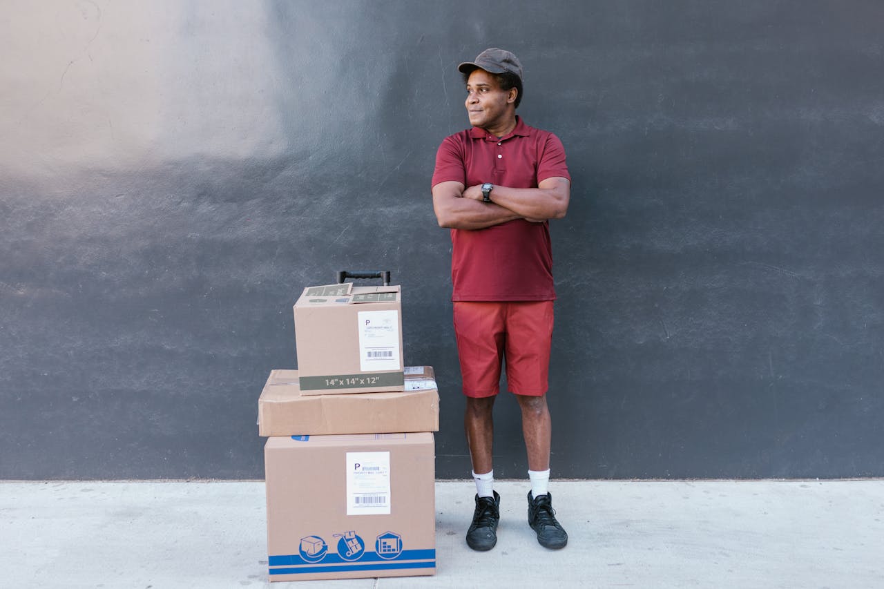 Delivery man in red attire posing outdoors with stacked cardboard boxes against a concrete wall.
